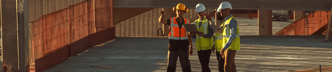 Three construction workers wearing hard hats and reflective vests look into the distance on a job site